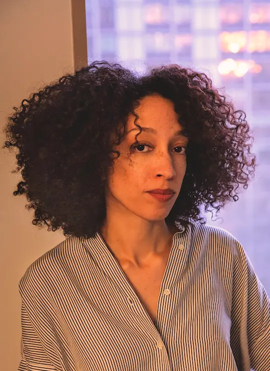 A Biracial Black woman
with coily, shoulder-length dark hair sits in front of a window.
She's wearing a striped blouse and looking at the camera,
slightly smiling.